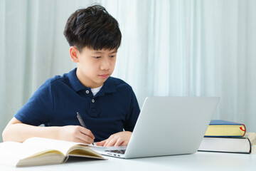 A smart and handsome asian teenager boy using notebook computer at the table to attend online classroom he look at screen and take notes while studying. Covid-19 New normal, Home Based Learning.