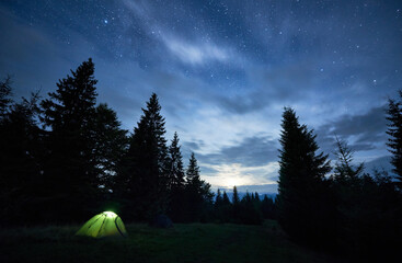 Wall Mural - Beautiful view of night starry sky over meadow. Stunning scenery of tourist illuminated green tent in forest with coniferous trees under blue sky with stars. Concept of travelling, hiking and camping.