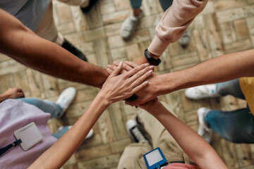 Top view of diverse young volunteers putting their hands on top of each other like a real team while standing in charitable organization office