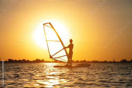 Plakaty Windsurfing  poczatkujaca-kobieta-windsurfer-stoi-na-desce-z-zaglem-na-tle-zachodu-slonca-windsurfing