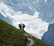 Couple of hikers travelling in Alps, man and woman standing on a trail passing through green meadow leading to breathtaking rocky mountains with snow