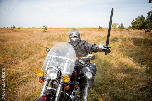 Fotomural A medieval knight in chainmail and a helmet with a sword in his hands sits on a motorcycle