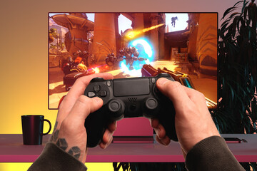 Wall Mural - Close up of Man Hands Holding Wireless Joysticks On Foreground. Modern TV Set With Shooter Video Game On Background. 3d rendering