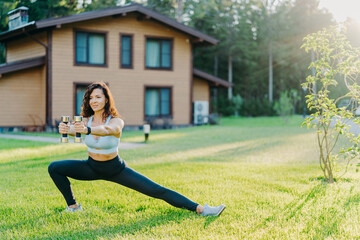 European sportswoman holds dumbbells and stretches legs, dressed in sport clothes, does physical exercises outdoor near house early in morning. Healthy lifestyle, weightlifting and fitness concept