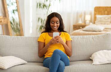 Wall Mural - Cheerful black lady sitting on cozy sofa with cellphone and messaging to her friend, indoors