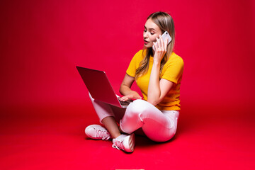 Happy young woman talking on the phone sitting on the floor with crossed legs and using laptop on red background.