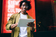 © BullRun - Cheerful handsome curly male in eyewear enjoying share and reading news on social networks via digital tablet, smiling 20s hipster guy standing outdoors near neon lights downloading application