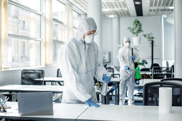 Disinfection of premises after work during covid-19 epidemic. People in hazmat suits, goggles and masks clean furniture with chemicals in office interior