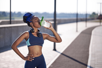 portrait of sporty african woman drinking water from bottle, sweaty female stand after running, outdoors