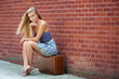 Stunning young blonde woman - in strapless sundress waits against brick wall sitting on vintage suitcase