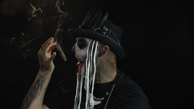 Scary guy in carnival costume of Halloween skeleton smoking cigar, making faces, showing tongue