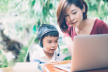 Wall Mural - Young asian mother and son using laptop computer for study and learning together at home, boy wearing headphone for e-learning with distancing, teacher or mom support child, education concept.