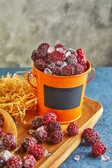 Frozen blackberries with ice in a metal orange bucket with a Mock up place on a wooden board and with hay on a blue background