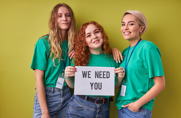 Wall Mural - beautiful caucasian women waiting for merciful volunteers in their organization, they need active altruistic people, wearing green t-shirts, isolated over green background