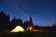 © anatoliy_gleb - Night camping near fire, forest and mountains on background. Group of friends warming up near bright bonfire. People sitting near tourist illuminated tent under night sky full of stars and milky way.