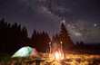 © anatoliy_gleb - Night summer camping in forest. Bright campfire burning, three male tourists standing around fire near tent under beautiful dark starry sky and Milky way. Concept of tourism, camping in the mountains.