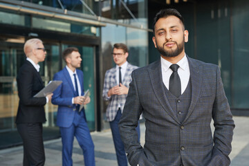 Wall Mural - Portrait of young leader in elegant suit looking at camera while standing outdoors with his team in the background