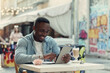 © gorynvd - Young happy african american man uses tablet pc sitting on street cafe with graffiti on backdrop.