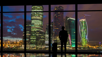 Wall Mural - The man with suitcase standing near a panoramic window on skyscrapers background