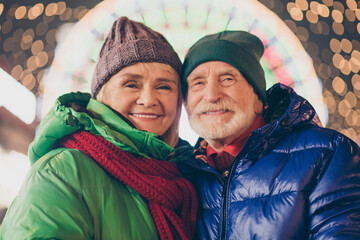 Wall Mural - Photo of two people retired lovers couple cozy cuddle cheek-to-cheek beaming smile amusement park newyear eve wear coat red scarf headwear x-mas evening street park lights fair outside