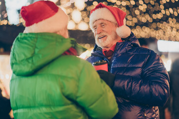 Wall Mural - Photo of two people pensioner man woman meeting hold cup drink mulled-wine cheerful smile retail choose decorations wear coat headwear mittens night street illumination fair outdoors