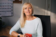 © Girts - Portrait of smiling mature woman in her home office. Working from home businesswoman. Adult european female entrepreneur looking at camera sitting at her desk. Close up portrait indoors