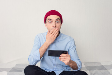 Surprised guy is sitting at home, young man is reading a text message on his phone, portrait