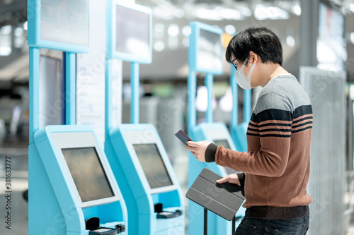 Asian man tourist wearing face mask using self check-in kiosk in airport terminal. Coronavirus (COVID-19) pandemic prevention when travel abroad. Health awareness and social distancing concept