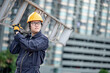 © zephyr_p - Asian maintenance worker man with protective suit and safety helmet carrying aluminium step ladder at construction site. Civil engineering, Architecture builder and building service concepts