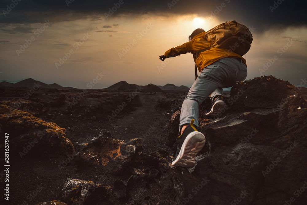 Successful hiker hiking a mountain pointing to the sunset. Wild man with backpack climbing a rock over the storm. Success, wanderlust and sport concept. - obrazy, fototapety, plakaty 