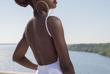 Fashion African Black Girl In A White Dress, Model Posing On A Background Of Blue Sky. Young African American Girl Model In White Dress With Open Back Posing Against Blue Sky