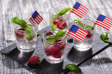 Alcohol shots of berries cocktail with a raspberry and basil on black stone tray. Glass of sparkling water with american flag on wooden table