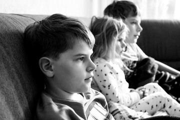 Cute little toddler girl and two school kid boys watching cartoons or movie on tv. Three happy healthy children, siblings during coronavirus quarantine staying at home. Brothers and sister together.