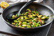 Roasted / fried brussels sprouts in a black frying pan sitting on a cutting board with a solid spoon for serving. Half a lemon and towel in the background