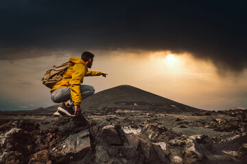 Wall Mural - Successful hiker hiking a mountain pointing to the sunset. Wild man with backpack climbing a rock over the storm. Success, wanderlust and sport concept.
