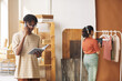 © AnnaStills - Young African man talking on the phone and working online on tablet pc he taking the order with woman working with clothes in the background