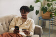 © AnnaStills - African smiling man lying on sofa with mobile phone and using credit card for buying or paying online