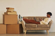 © AnnaStills - African young man lying on sofa with laptop and working with online orders with big stacks of parcels on the floor prepared for shipping