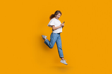 Emotional caucasian blonde woman running and jumping on a yellow studio wall while chatting on mobile and promoting something