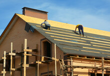Roofing Contractors Are Installing Underlayment, Waterproofing Membrane On A Roof Deck, Plywood Sheathing Of A Large Brick House Under Construction With Scaffolding.