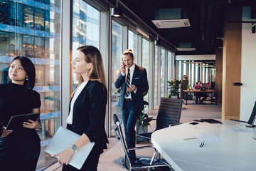 Wall Mural - Confident male entrepreneur in formal wear talking on mobile phone walking in office while his female colleagues having conversation,businessman making smartphone call during working process