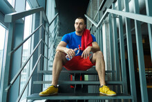 Bearded Muscular Man Wears Blue T-shirt, Yellow Sneakers And Red Shorts Drink Water From Blue Bottle In The Gym.