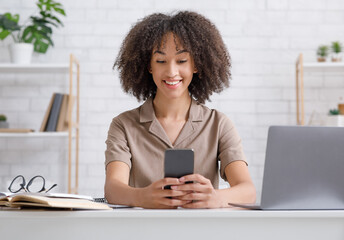 Wall Mural - Modern technology for study and work at home during epidemic. Smiling african american woman typing on smartphone, sitting at table with notebook and notepad