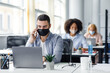 © Prostock-studio - Customer consultation by phone remotely at returning to work after quarantine. Millennial man in protective mask with smartphone looks at laptop at workplace