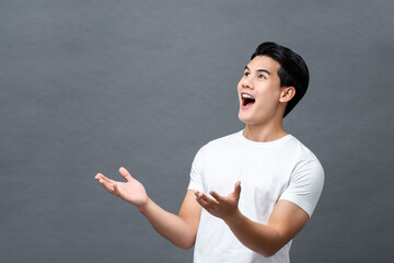 Portrait of surprised smiling young handsome Asian man looking upward and presenting with open palms gesture in isolated studio gray background