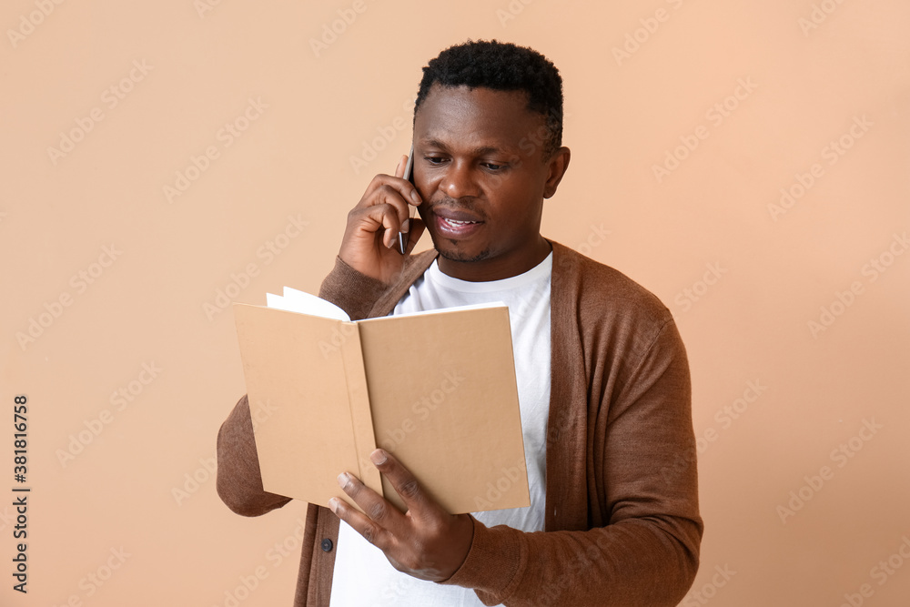 African-American man with book talking by phone on color background