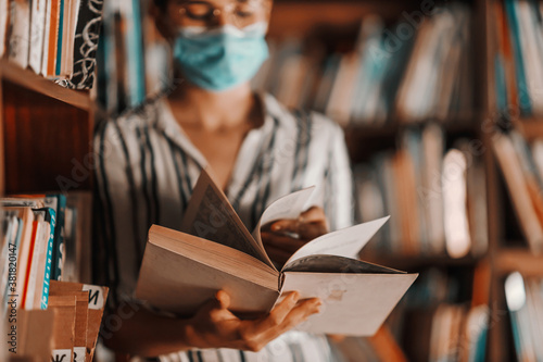 Closeup of attractive college girl standing in library with face mask on and reading a book. Studying during corona virus concept.