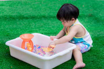 Asian boy are using toys to scoop colorful water beads. Son learns his senses through play. Children sit on the green lawn. Sweating on your face in hot weather. Age 2 years old.