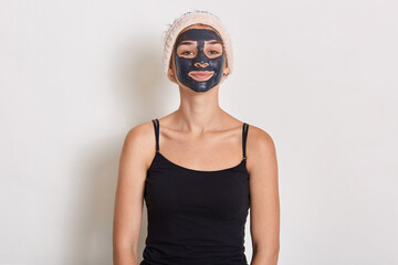 Spa teen girl applying black facial clay mask. Beauty treatments at home. Lady in sleeveless t shirt and hair band looking directly at camera, posing isolated over white background.