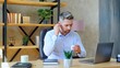 © viacheslav - Businessman sitting on chair at table in home office puts on wireless earphones using laptop computer. Gray-haired bearded man connects to remote online video chat conference with company employees